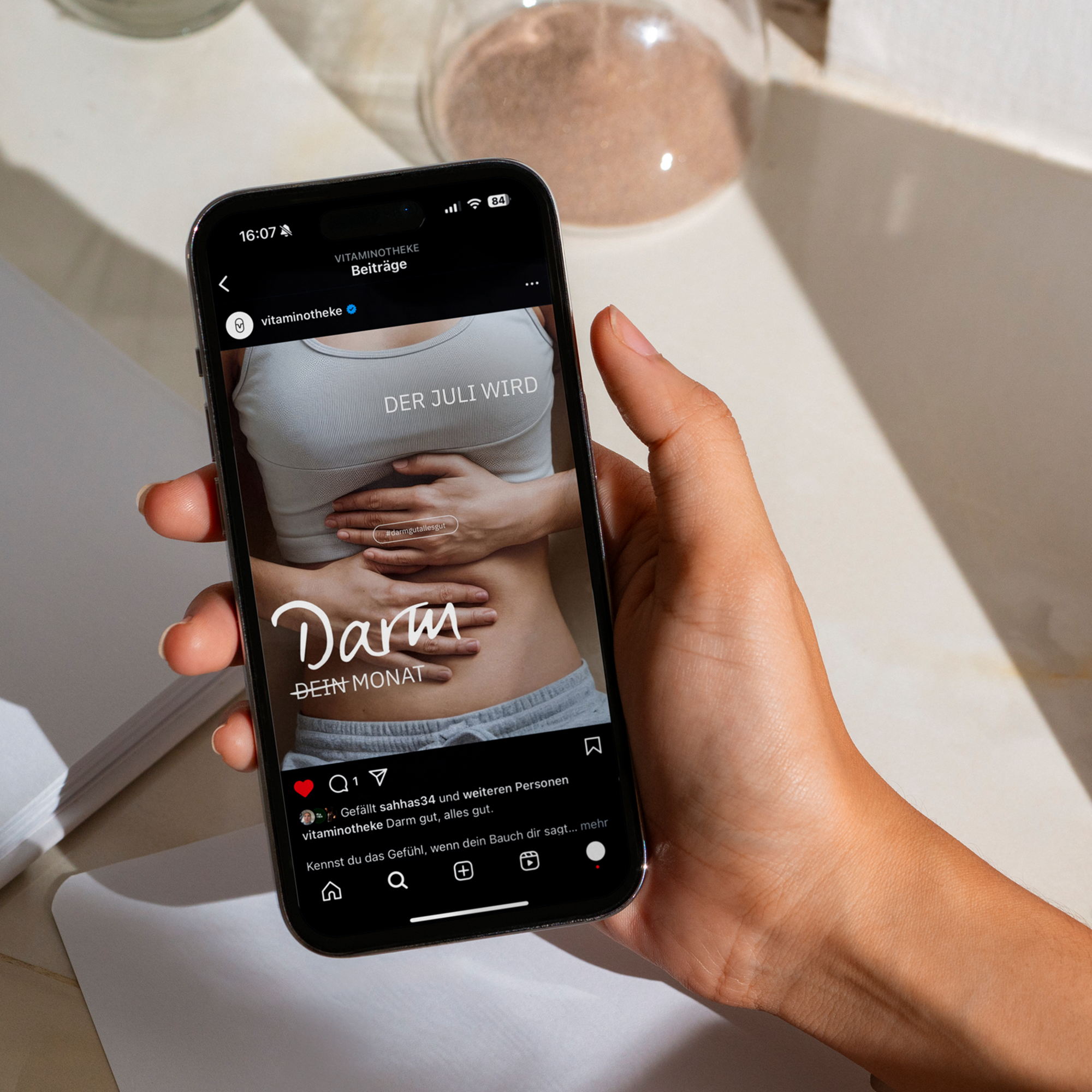 Eine Hand, die ein Handy hebt. Auf dem Bildschirm ist ein Instagram Post von der Vitaminotheke zu sehen. Darauf steht Der Juli wird dein und Darm Monat.